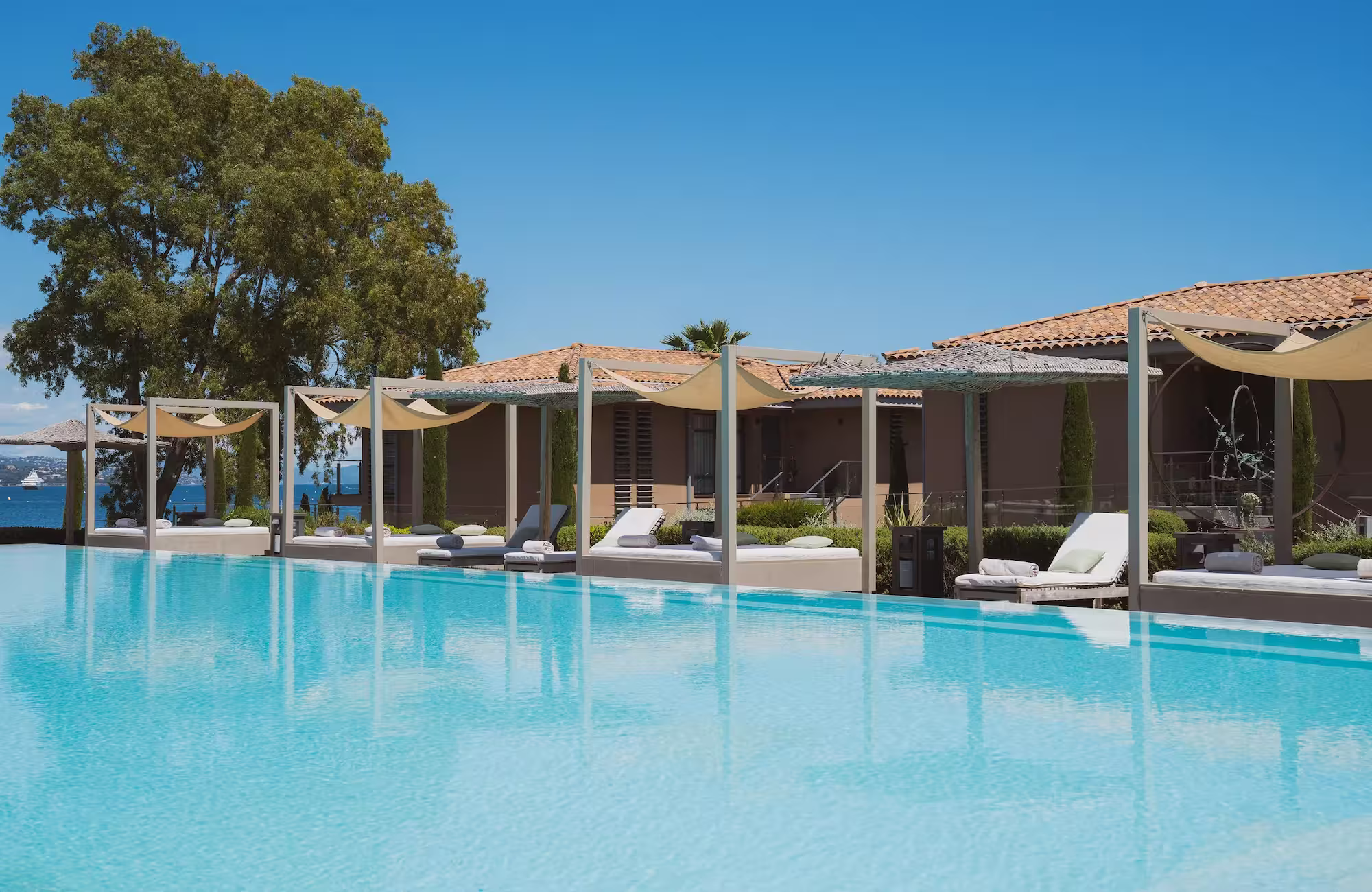 Piscine à débordement avec des chaises longues sous des pergolas près d'une maison à toit en tuiles.