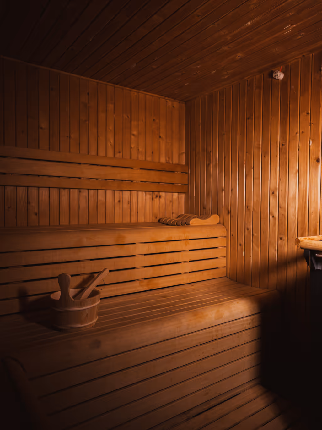 Intérieur d'un sauna en bois avec des bancs, un seau et une louche en bois, éclairage tamisé.