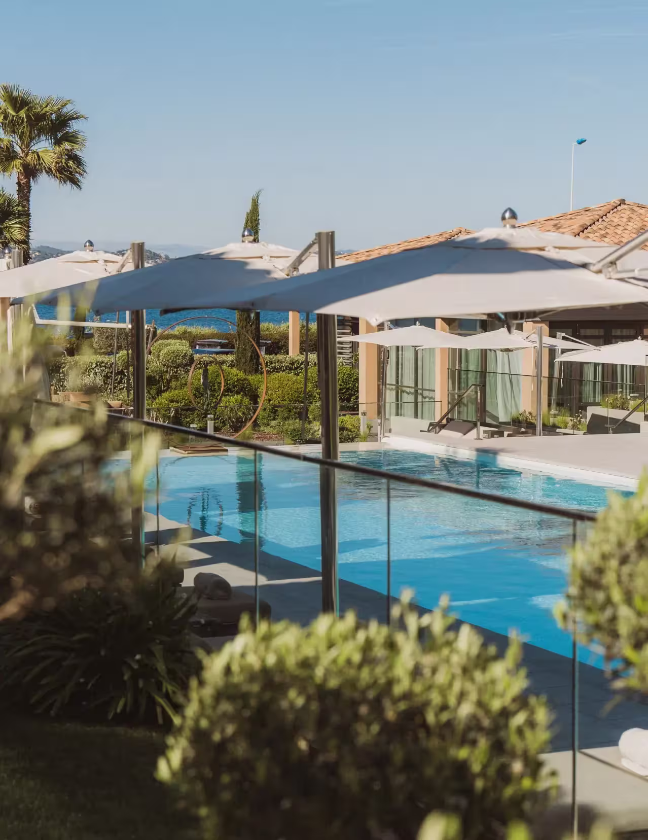 Piscine extérieure avec parasols blancs et plantes, vue sur un bâtiment moderne avec toits en tuiles sous un ciel bleu.