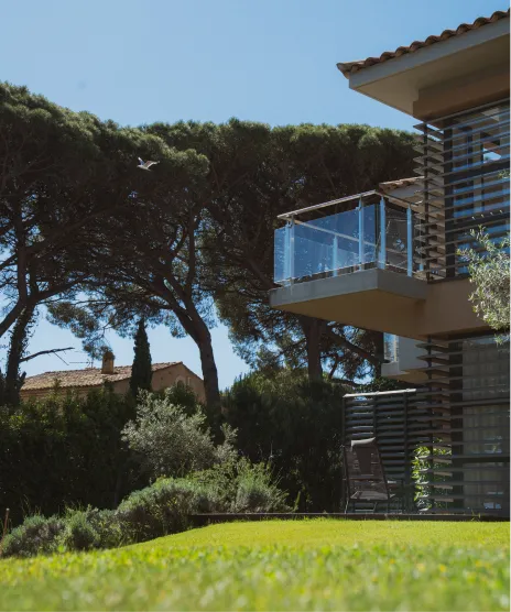 Vue d'une maison moderne avec un balcon en verre sur un jardin vert, arbres et ciel bleu en arrière-plan.