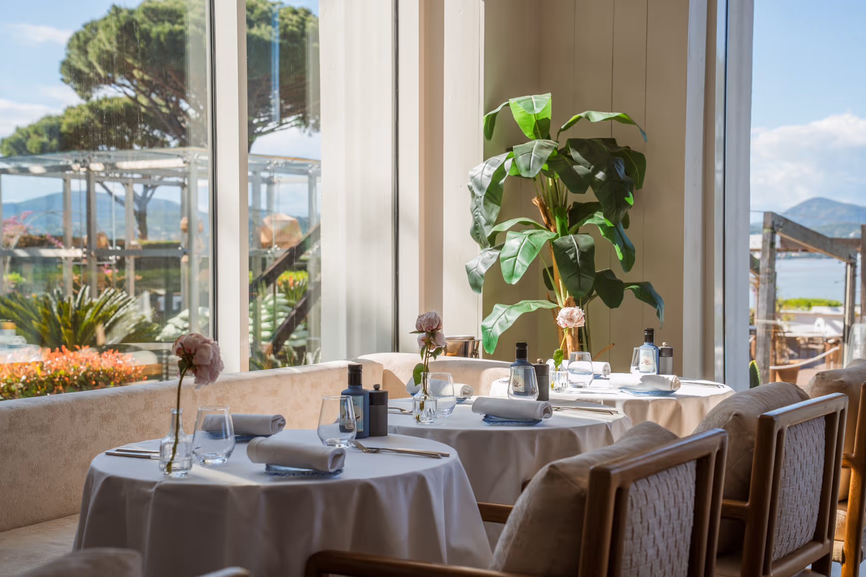 Tables élégamment dressées dans un restaurant lumineux avec grandes fenêtres offrant une vue sur un jardin et la mer.