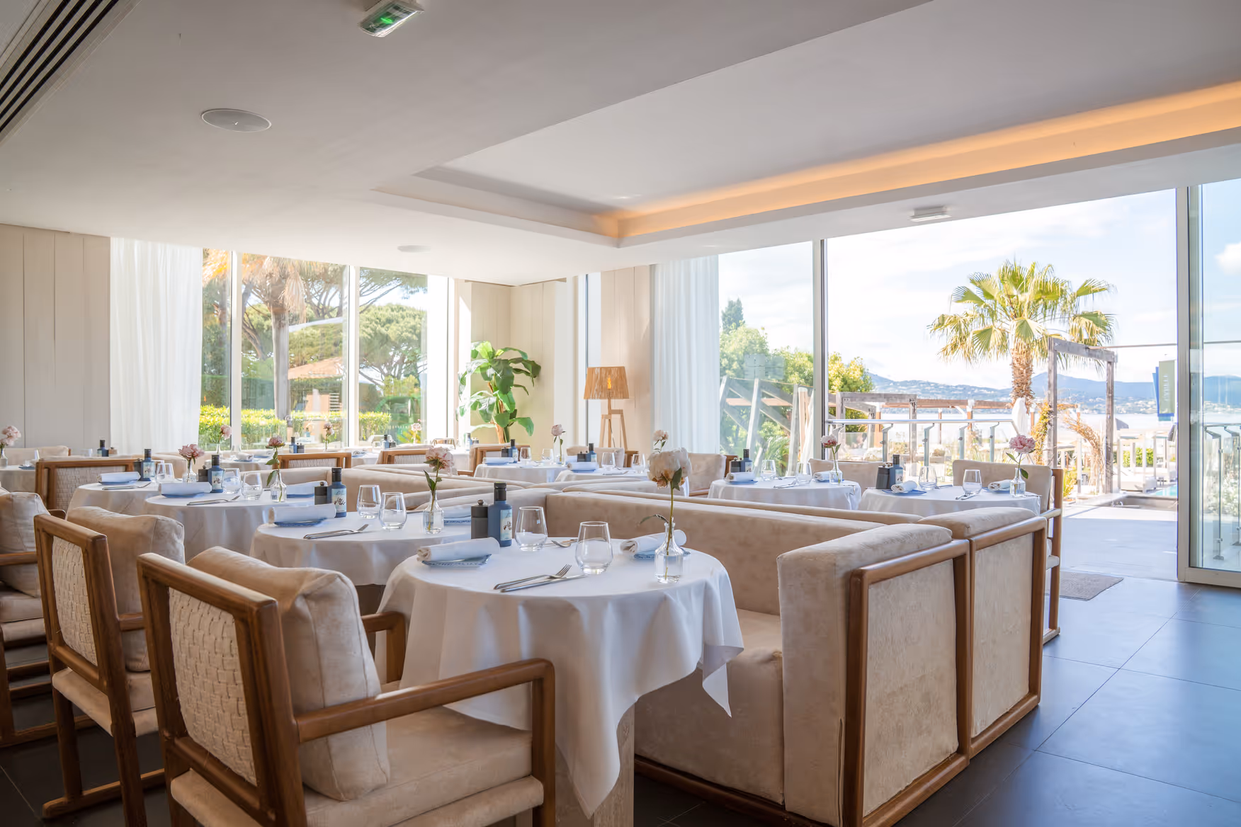Salle de restaurant du Kube Saint-Tropez élégante avec tables rondes dressées de nappes blanches, chaises en bois et canapés, grandes baies vitrées offrant une vue sur des palmiers et la mer.