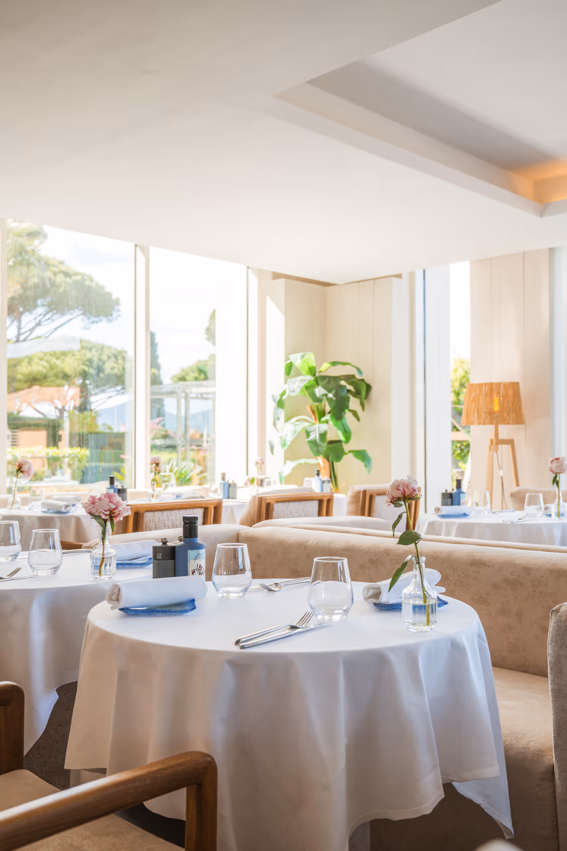 Table ronde élégamment dressée avec nappe blanche, serviettes, verres, couverts, et fleurs roses dans un restaurant du Kube Saint-Tropez lumineux avec de grandes fenêtres.
