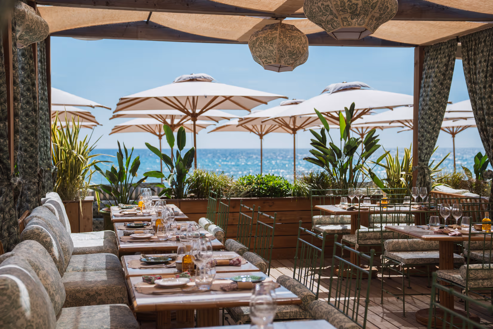 Terrasse de restaurant de la plage Cybele en bord de mer avec tables dressées, chaises et parasols blancs, vue sur l'océan sous un ciel bleu clair.