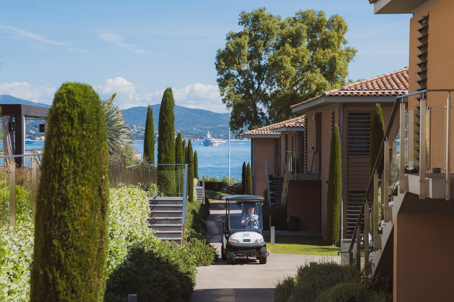 Voiturette de golf conduite entre des bâtiments modernes avec des arbres et une vue sur la mer et un yacht à l'arrière-plan.