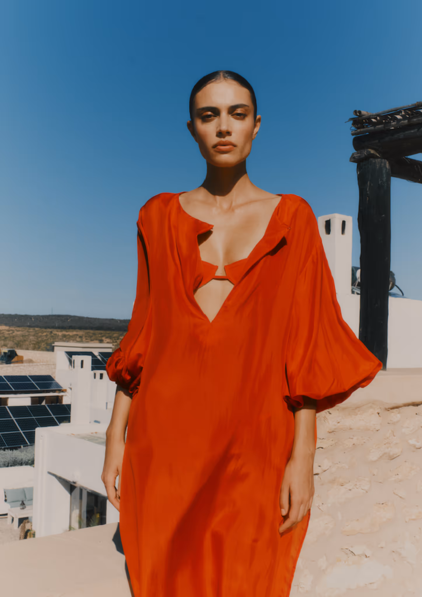 Femme portant une robe rouge fluide avec des manches bouffantes, posant dehors sous un ciel bleu clair.