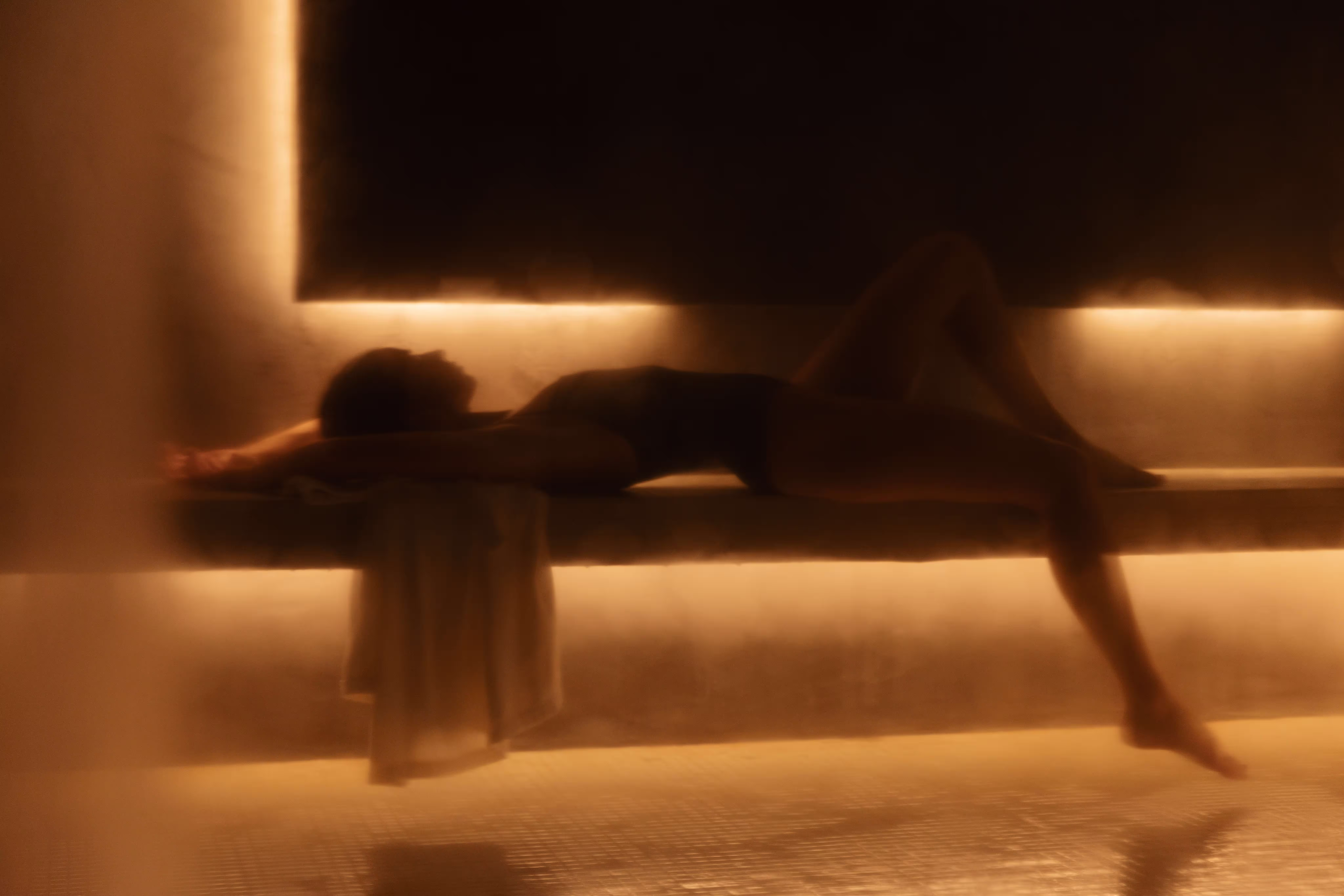 Personne allongée dans un sauna chaud avec des lumières tamisées et une serviette suspendue.