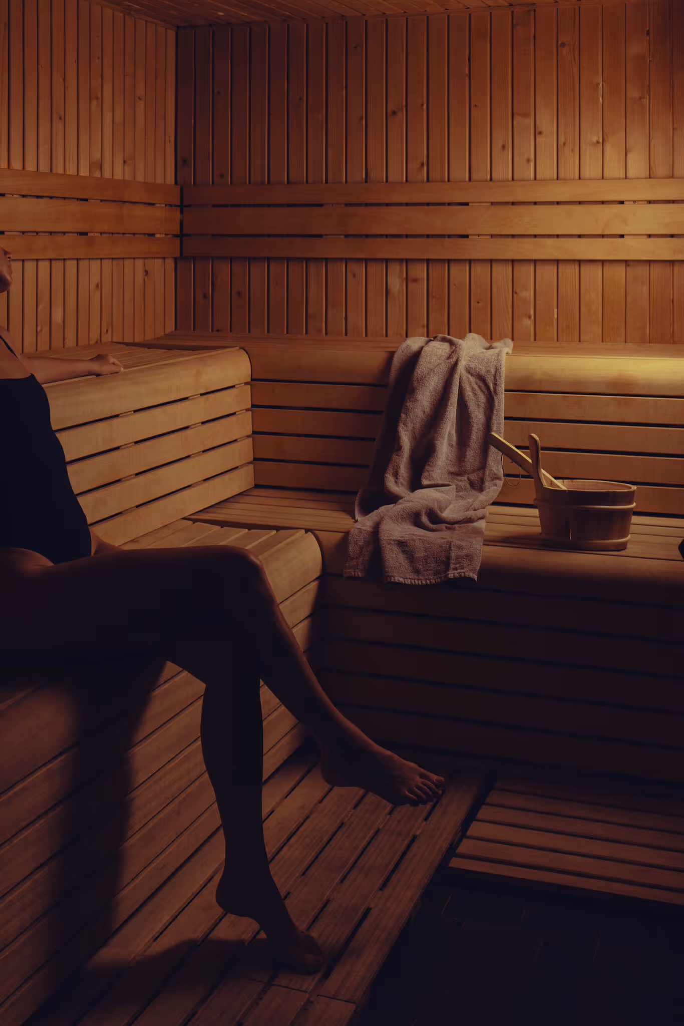 Personne assise dans un sauna en bois avec une serviette et un seau posés sur la banquette.