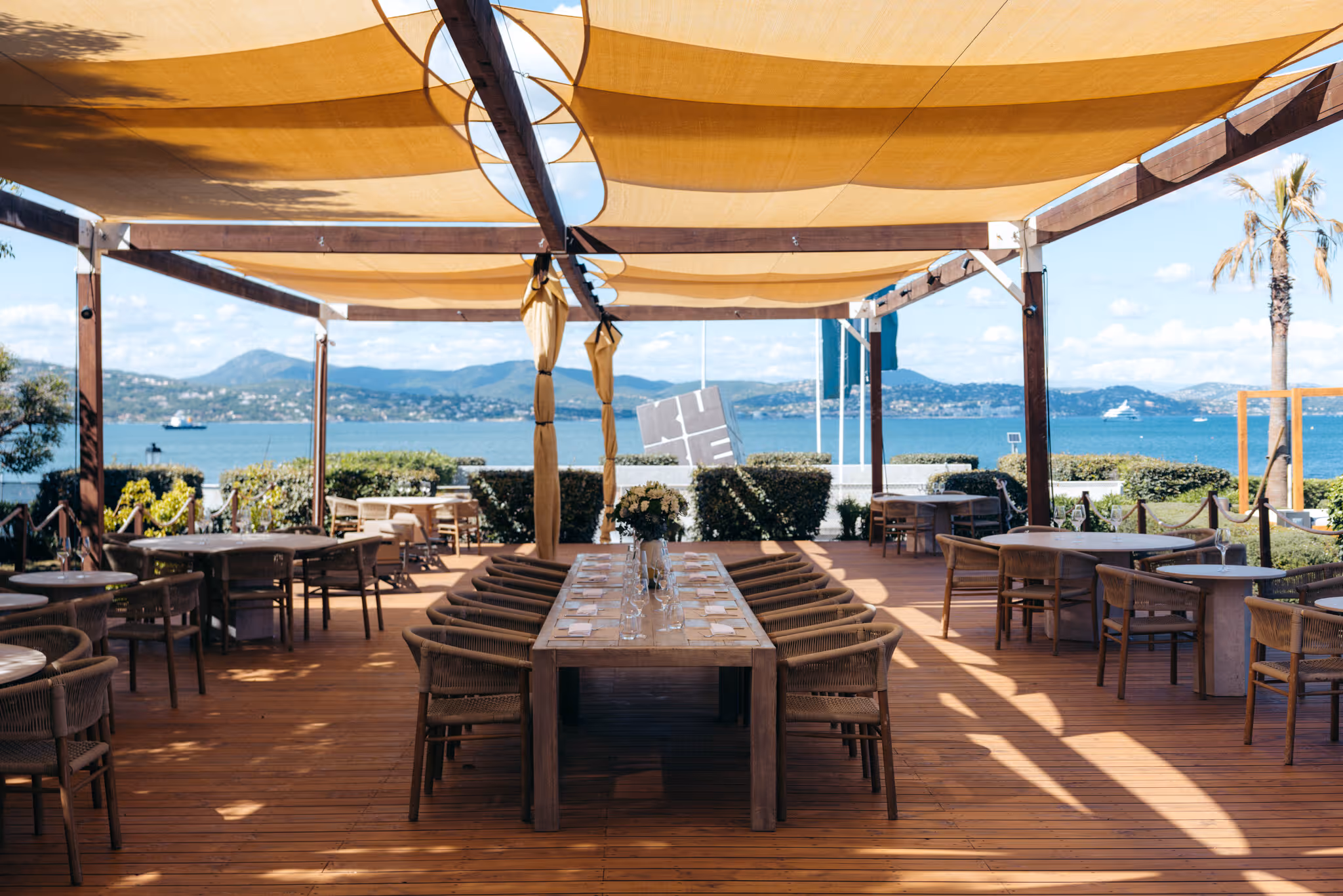 Terrasse de restaurant avec table en bois longue, chaises, vue mer et montagnes, ombragée par voiles jaunes.