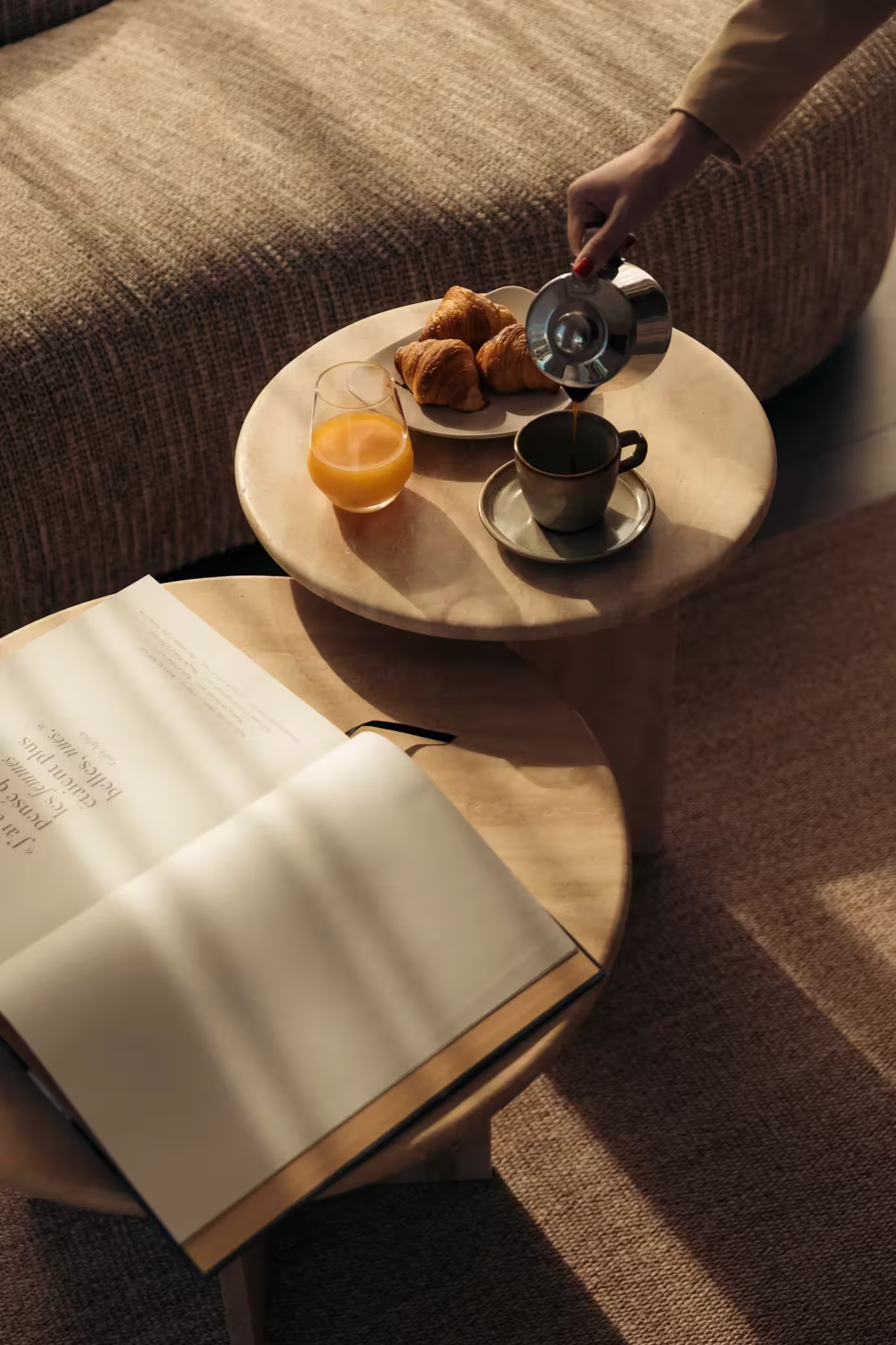 Main vers versant du café dans une tasse avec jus d'orange, croissants et livre ouvert sur la table.