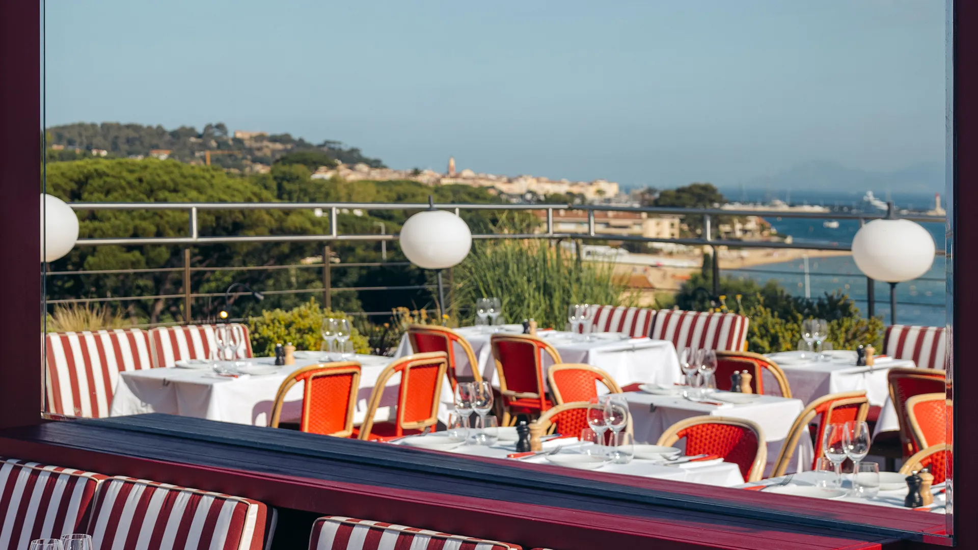Terrasse de restaurant avec tables dressées, chaises rouges, vue sur la mer et la ville au loin.