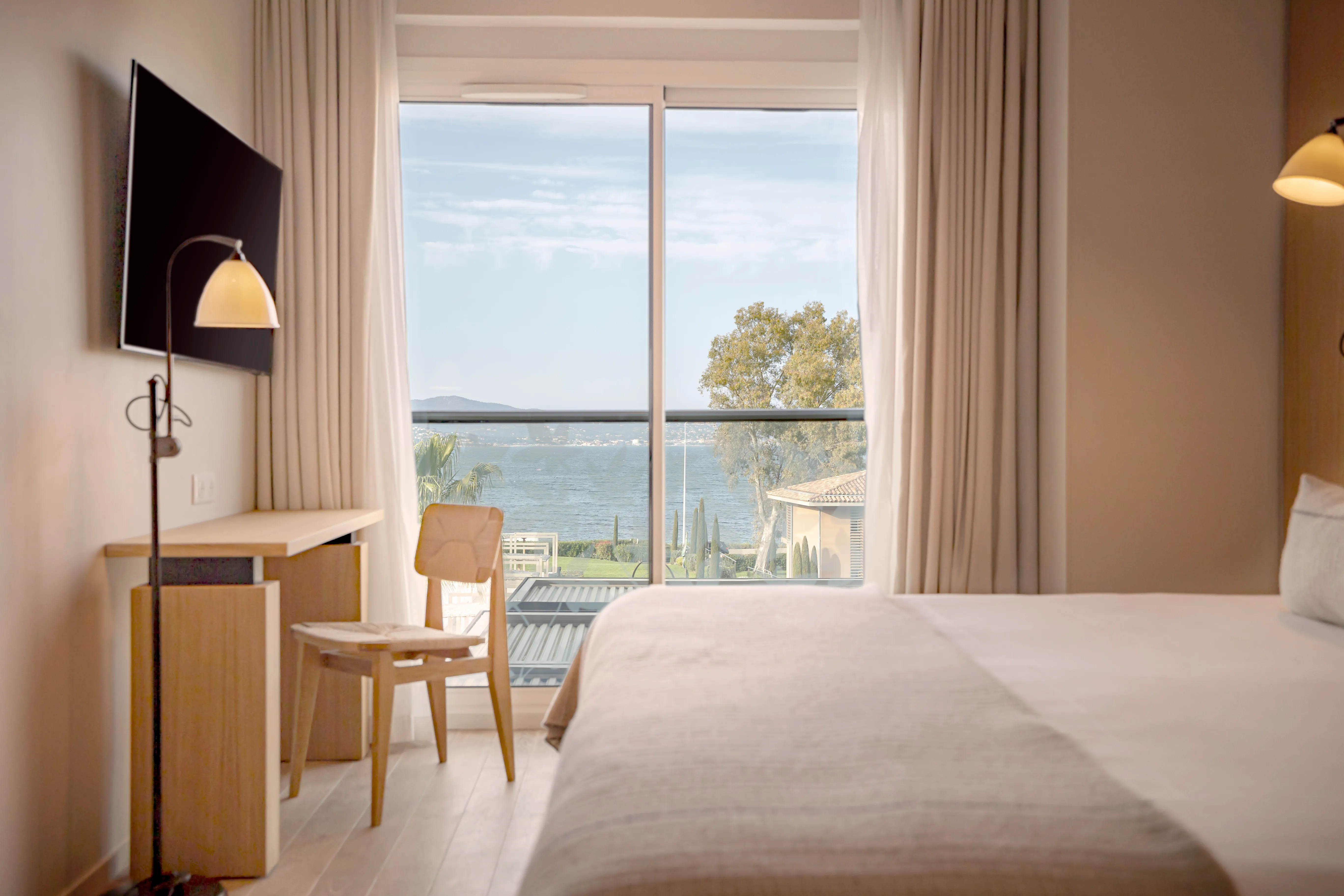 Chambre lumineuse au design naturel avec vue sur mer, rideaux clairs en lin et mobilier en bois chaleureux.